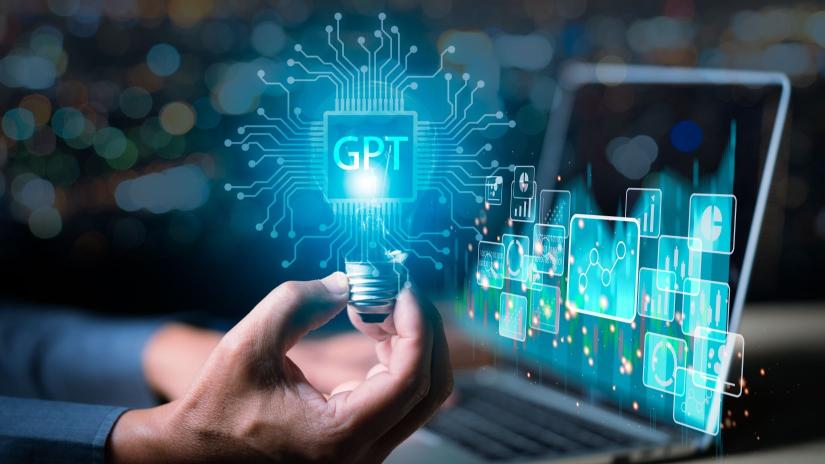 Close up on hand holding a lightbulb with a hologram with letters "GPT" with laptop in the background. 