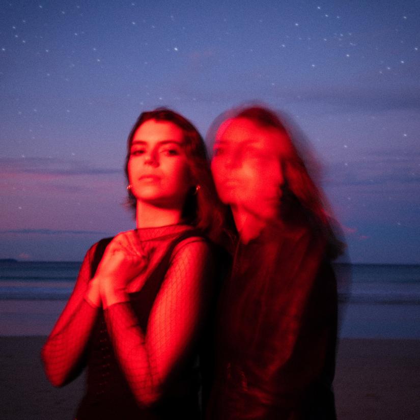 Image of two females standing next to each other in red lighting at the beach at night time. The image is deliberately blurry and moody.