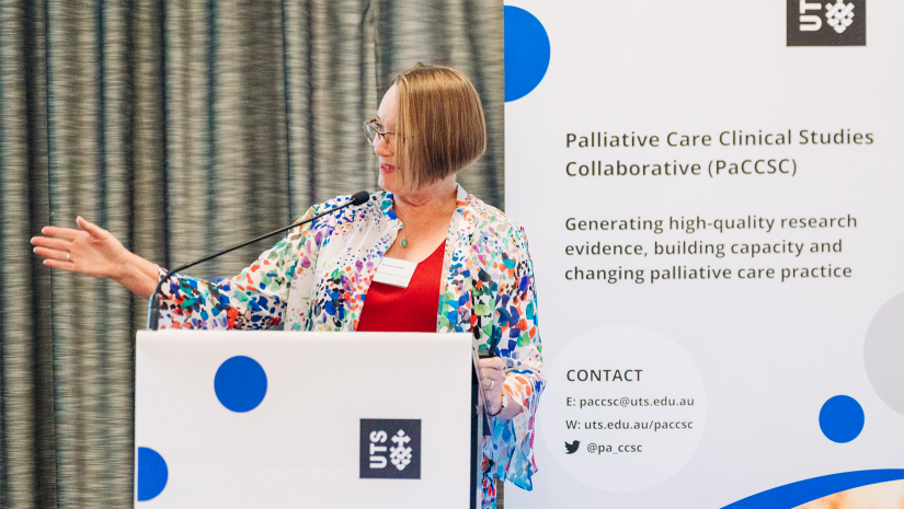 Belinda Fazekas presenting at a lectern. she is standing in front of a PaCCSC promotional banner, which has a grey background and blue spots. Belinda is wearing a red top under a floral cardigan. 