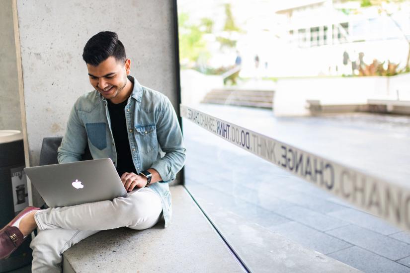 male students sitting by window pane on laptop wearing a blue jean jacket and white pants