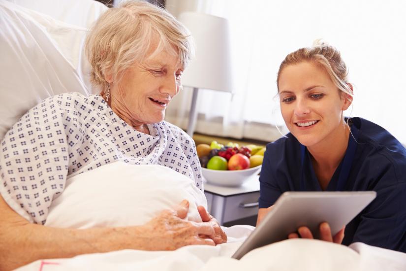 Young woman talking to elderly woman who is in bed looking at a tablet.