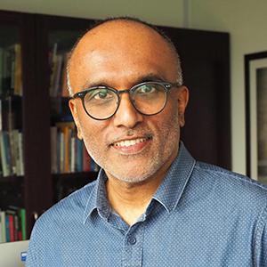 Cherian smiles at the camera, wearing glasses and a blue button-up shirt in front of a wall of books