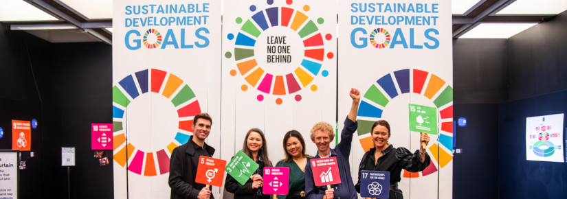 Five members of the UTS BUILD and Sustainability programs stand smiling in front of three vertical banners that read “SUSTAINABLE DEVELOPMENT GOALS”. The banner features multicoloured circular graphics. The members hold orange, green, pink and blue signs that correlate with the United Nations 17 Sustainable Development Goals.