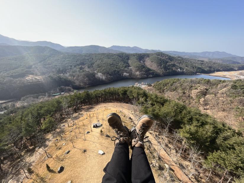 Photo of Meg Brown's feet in hiking boots sticking out in front of a view of trees, mountains and a river or lake.
