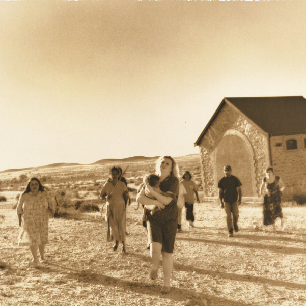 A sepia toned photograph of a woman carrying a baby, running away from a group of people