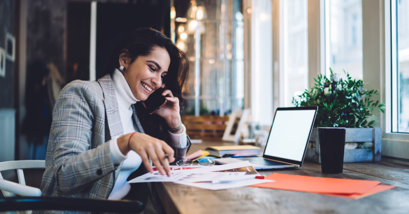 A women in a grey jacket smiles as she talks on the phone and looks down at paper reports and her laptop. 