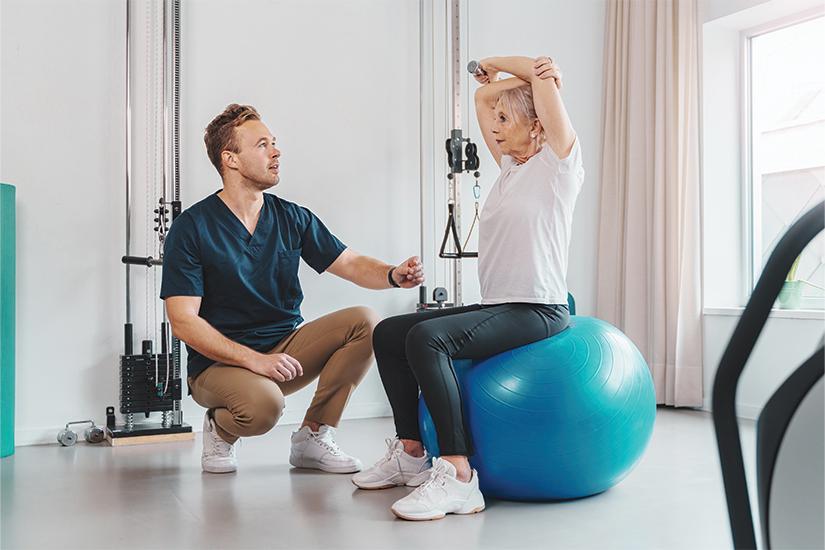 Elderly woman sitting on an exercise ball with a physio kneeling next to her instructing her.