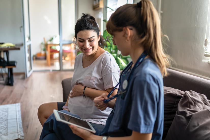 Pregnant woman holding her belly looking down at tablet being held by midwife.