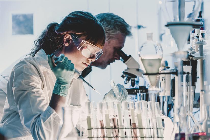 Female research in the foreground wearing safety goggles and a research lab coat. She is holding a syringe and putting a liquid into a test tube.  Male researcher in background looking into a microscope.