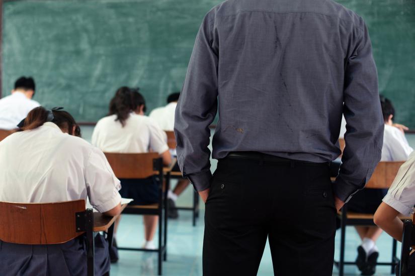 Stock picture of a teacher supervising an exam in a classroom standing behing a group of students seated at desks.