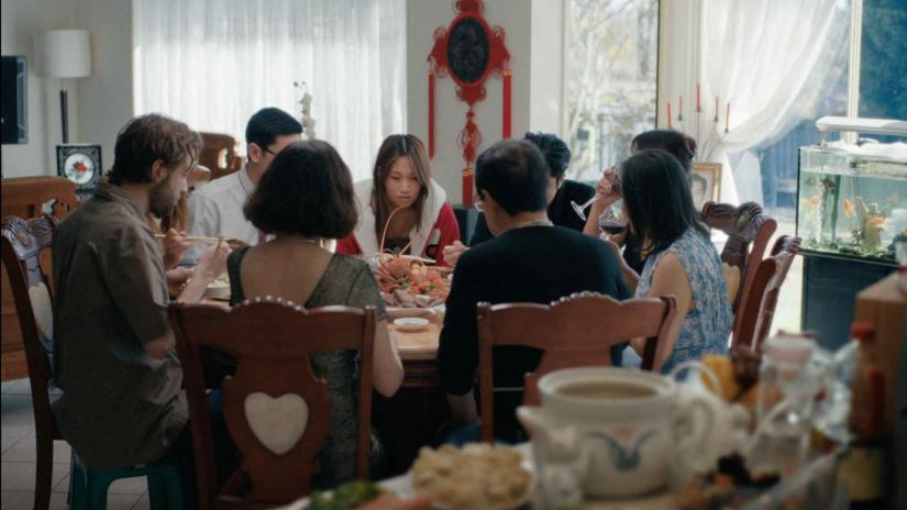 A family sits around a table during a Lunar New Year celebration. The view is focused on Mary, a young woman who sits staring at her plate while those around her talk and eat.