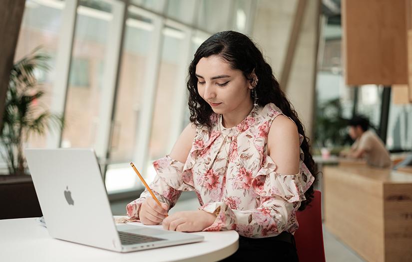 A young woman sitting at a desk with a laptop, holding a pencil to write on some paper 