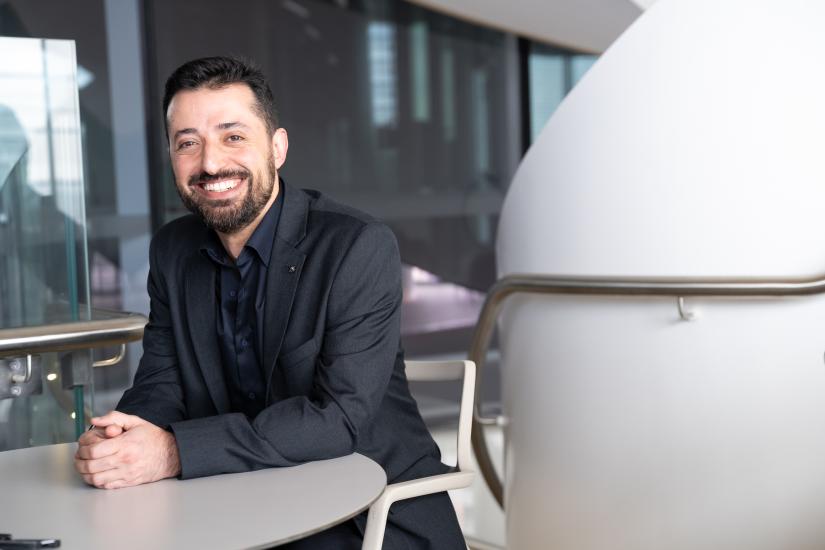 Image of man with dark hair and beard wearing black jacket, sitting, smiling