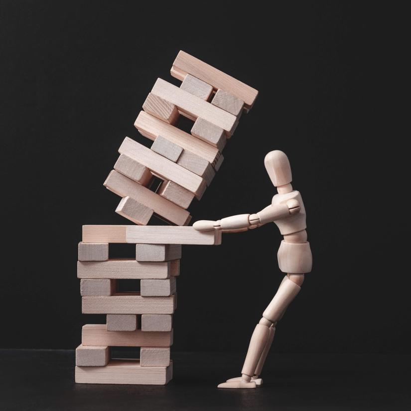 A figure pulls a piece from a jenga puzzle that towers over them and is about to topple