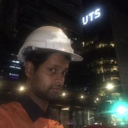 Nightime shot of man wearing a hard hat facing right in front of a skyscraper that has UTS lit up in lights.