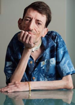 Man looking at the camera with his chin resting on his hand with a blue button up shirt and two gold bracelets