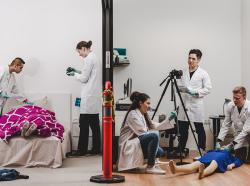On the left side of a room, two forensic students are examining a mannequin covered by a blanket on a bed. On the right side, there are three students. Two are examining a mannequin on the floor while the third student is standing in front of a camera. 