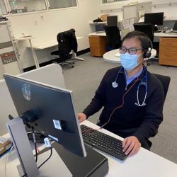 Dr Wei Lee wearing glasses, surgical mask and a navy jumper sits at a computer for a telehealth consultation. He is wearing headphones and has a stethoscope around his neck.