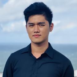 Man standing in front of ocean and wearing a black collared shirt looks into camera.