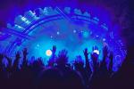A music stage illuminated in blue lights and haze with shadows of people in the crowd with their hands up