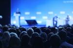 Blue toned image of participants at a conference and blurred stage with presenter