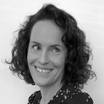 Black and white photo of Megan Taylor smiling at the camera, taken from the shoulders up. Megan has shoulder-length hair that is curly and dark. She is wearing a dark-coloured top with small light-coloured shapes dotted throughout.